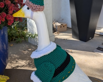 May include: A white goose ornament dressed in a green and black crochet sweater with white trim and an elf hat. The hat is green, yellow, and red. The goose is on a green base.