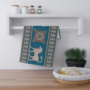 May include: A teal kitchen towel with a brown and white geometric pattern featuring a lion and a circular design. The towel is hanging on a white shelf.