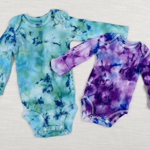 Op de afbeelding: Twee baby-rompers met lange mouwen met tie-dye patronen. Eén romper is blauw, groen en wit. De andere romper is paars, blauw en wit.