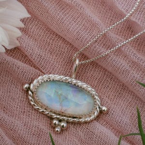 May include: A silver necklace with an oval pendant featuring a shimmering, iridescent stone. The pendant is framed by a twisted rope design and small silver beads. The necklace rests on a pink fabric, with white flowers and green foliage in the background.