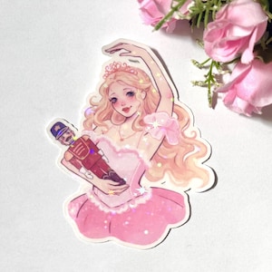 May include: A sticker featuring a ballerina in a pink tutu holding a toy soldier. The ballerina has blonde hair and is wearing a crown. The sticker has a glittery finish.