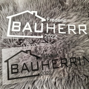 Könnte beinhalten: Zwei transparente Vinyl-Aufkleber mit einem Haussymbol und dem Text "Bauherr" in Weiß und Schwarz. Der obere Aufkleber trägt den Text "Frederic Bauherr 2022" und der untere Aufkleber trägt den Text "Stefanie Bauherrin 2022".