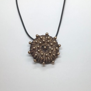 May include: A gold-toned pendant necklace with a round, textured design resembling a sea urchin. The pendant is made of wire and small beads, and is suspended from a black cord.