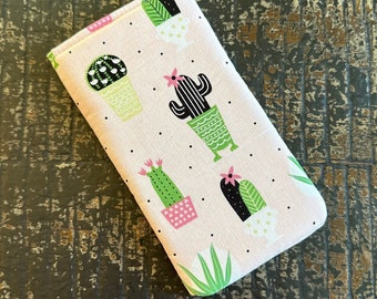 May include: A rectangular, light pink fabric case with a repeating pattern of cacti in various pots and styles. The cacti are green, black, and white with pink flowers. Black dots are scattered across the background.
