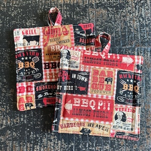 May include: Two square BBQ-themed pot holders with red and black checkered patterns. The fabric features various BBQ-related text and graphics, including "BBQ," "Smoke Out Here," and "Barbecue Heaven." Each pot holder has a red and white checkered loop for hanging.
