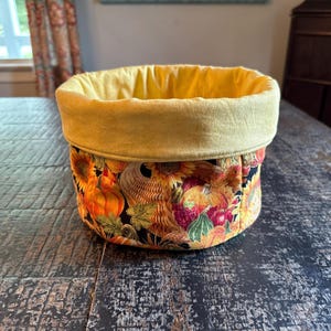 May include: A fabric basket with a yellow interior and a fall-themed exterior. The exterior features pumpkins, sunflowers, and a basket design on a black background. The basket is sitting on a dark wooden surface.