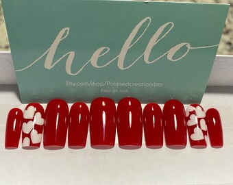 May include: Set of ten red press-on nails with white heart designs. The nails are oval shaped and have a glossy finish. Etsy.com/shop/Polishedcreationsbar Press on nails.