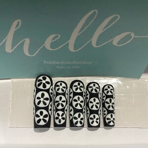 May include: Five black press-on nails with a white floral design. The nails are long and have a rounded shape. The design features white flowers with black centers on a black background. The text "Polishedcreationsbar Press on nails" is visible in the background.