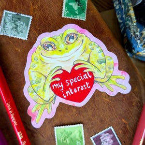 Op de afbeelding: Een grillige sticker met een gele kikker die een rood hart vasthoudt met de tekst "my special interest". Vintage postzegels en kleurpotloden omringen de sticker op een bruin leren oppervlak.