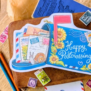 Puede incluir: Una pegatina troquelada con forma de buzón azul lleno de sobres y un caracol. El buzón presenta girasoles amarillos, una bandera roja y el texto cursivo blanco Happy Postcrossing. Varios sellos postales están esparcidos por el diseño.