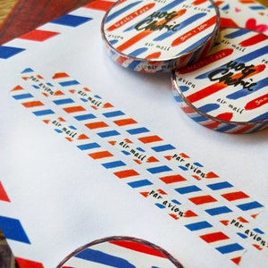 May include: Red, white, and blue washi tape with the text "air mail" and "par avion" printed on it. The tape is rolled up and has a silver backing. The tape is on a white surface.