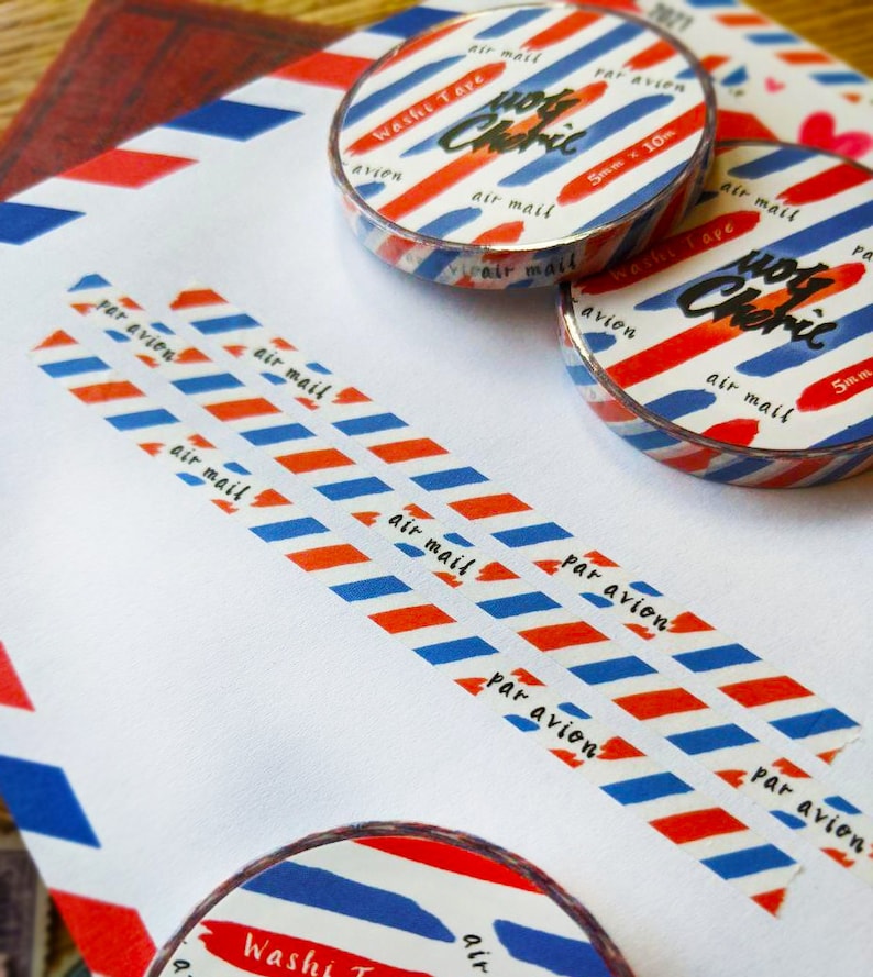 May include: Red, white, and blue washi tape with the text "air mail" and "par avion" printed on it. The tape is rolled up and has a silver backing. The tape is on a white surface.