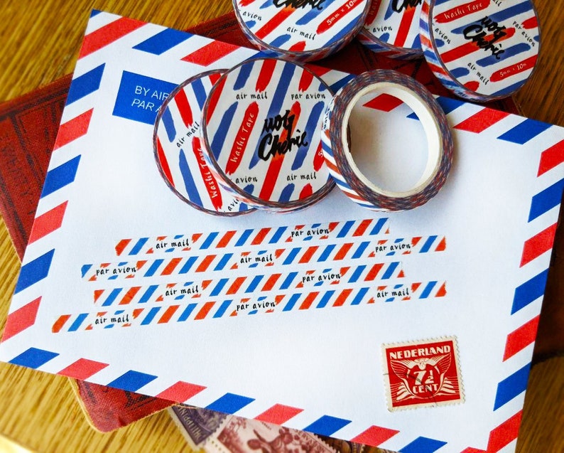 May include: Red, white, and blue washi tape with the text "Washi Tape" and "Air Mail" in black. The tape is on a white envelope with a red, white, and blue striped pattern. The envelope also has a stamp with the text "NEDERLAND" and "7 CENT" in black.