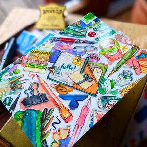 Puede incluir: Una tarjeta blanca con una ilustraci&oacute;n colorida de varios art&iacute;culos de papeler&iacute;a, incluyendo bol&iacute;grafos, l&aacute;pices, tijeras, cinta adhesiva y una libreta con la palabra "&iexcl;hola!" escrita en ella.