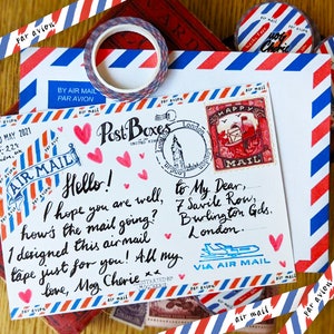 May include: A white envelope with red and blue stripes, decorated with washi tape and hearts. The envelope has a stamp with the words "Happy Mail" and a handwritten message that reads "Hello! I hope you are well, how's the mail going? I designed this airmail tape just for you! All my love, Mag Cherie."