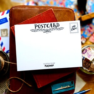 Puede incluir: Una postal blanca en blanco con las palabras "POSTCARD" impresas en negro en la parte superior. La postal est&aacute; encima de un libro encuadernado en cuero marr&oacute;n. La postal tiene un peque&ntilde;o sello en la esquina superior derecha que dice "very Cherished".