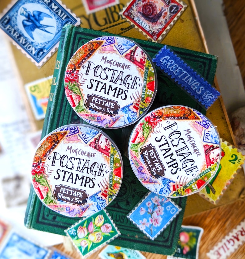 Puede incluir: Tres rollos de cinta washi colorida con un dise&ntilde;o de sello de correos vintage. La cinta est&aacute; etiquetada "Mogcherie Postage Stamps Pettape 50mm x 5m".