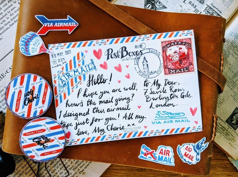 May include: A colorful airmail envelope with red, white, and blue stripes. The envelope has a handwritten message that says "Hello! I hope you are well, how's the mail going? I designed this airmail tape just for you! All my love, Mag Cherie." The envelope also has a stamp that says "Happy Mail" and a sticker that says "Via Air Mail".