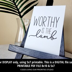 May include: A white printable sign with black text that reads "WORTHY IS THE lamb". The sign is displayed on a small wooden easel.