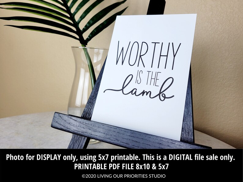 May include: A white printable sign with black text that reads "WORTHY IS THE lamb". The sign is displayed on a small wooden easel.