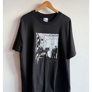 Puede incluir: Camiseta negra con una impresión blanca de una banda llamada "The Echos" con el texto "Repeat Performances Beginning March 18, 1998".