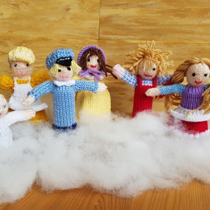 Może przedstawiać: Zestaw siedmiu dzianych palczkowych lalek, każda z innym wzorem i kolorem. Lalki są ułożone na białym, puszystym podłożu, przypominającym chmurę. Lalki obejmują białego anioła, postać ubraną na żółto i biało, postać ubraną na niebiesko i biało, postać ubraną na żółto i biało, postać ubraną na czerwono i biało, postać ubraną na fioletowo i biało oraz postać ubraną na czerwono i biało z blond włosami.