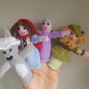 May include: Hand-knitted finger puppets depicting characters from a fairy tale. The puppets include a wolf, a girl in a red hood, an elderly woman, and a man in a green hat. Each puppet has a unique design and colour scheme.