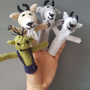 Peut inclure: Quatre marionnettes à doigts en tricot, deux sont des chèvres avec des barbes blanches, une est un monstre vert avec un bavoir bleu et une est une chèvre avec une barbe blanche. Les marionnettes sont sur une main.