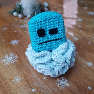 Può includere: Un personaggio blu all'uncinetto con un'espressione accigliata, che indossa un colletto bianco all'uncinetto. Il personaggio è seduto su una superficie di legno con un motivo a fiocco di neve.