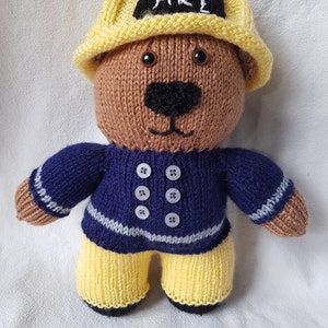 May include: A knitted brown teddy bear wearing a yellow firefighter hat with the word "FIRE" embroidered on it. The bear is wearing a blue sweater with white buttons and yellow pants.