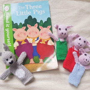 May include: A set of finger puppets featuring three pink pigs and a grey bunny. The puppets are made of knitted fabric and have embroidered details. The puppets are arranged on a white surface next to a children's book titled "The Three Little Pigs".