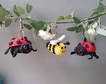 May include: Three crocheted keychains hanging from a branch. The keychains are a ladybug, a bee, and another ladybug. The ladybug keychains are red with black spots and black legs. The bee keychain is yellow and black with white wings.