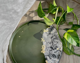Puede incluir: Una bandeja redonda verde con una bandeja de mármol blanco y negro en forma de semicírculo con detalles dorados en la parte superior. La bandeja está sobre una superficie de madera con una planta verde.