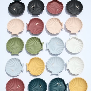 May include: Assortment of 18 decorative seashell-shaped dishes in various colors, including black, brown, beige, green, yellow, blue, and white. Each dish has a subtle gold fleck finish.