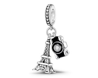 May include: A silver charm with a travel theme. The charm features a miniature Eiffel Tower and a black camera with a clear gem. The charm is suspended from a silver ring with heart-shaped details.
