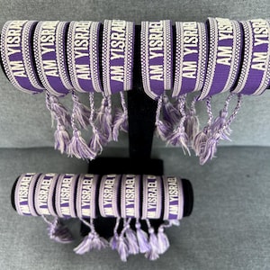 May include: A display of purple and white woven bracelets. Each bracelet features the text "AM YISRAEL" in white lettering. The bracelets are adorned with matching tassels and are displayed on a black stand.