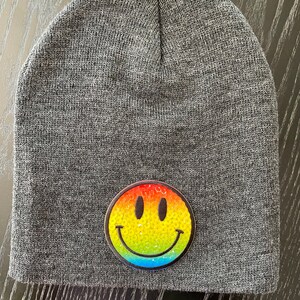 Puede incluir: Gorro gris con un parche de cara sonriente de lentejuelas arcoíris.