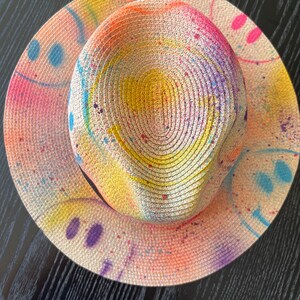 May include: A straw hat with a colorful paint splatter design. The hat is white with pink, yellow, blue, and purple paint splatters. The hat has a wide brim and a round crown.