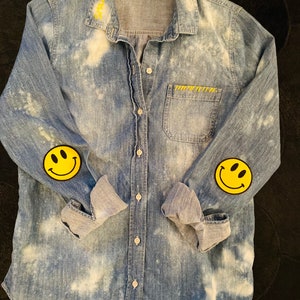 May include: A light blue denim shirt with a white bleach design. The shirt has two yellow smiley face patches on the sleeves.