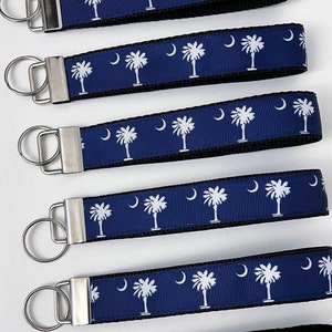 May include: Six blue and white keychains with a silver key ring. Each keychain features a repeating pattern of the South Carolina state flag, which depicts a palmetto tree and a crescent moon.
