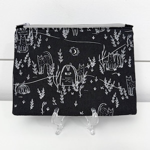 May include: Black zippered pouch with a white Halloween-themed design. The design includes cats, pumpkins, a tombstone with "R.I.P.", and a crescent moon. The pouch is rectangular and has a silver zipper.