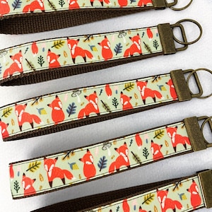 May include: Five brown keychains with a light green background and a repeating pattern of orange foxes and leaves. Each keychain has a gold-toned metal key ring.