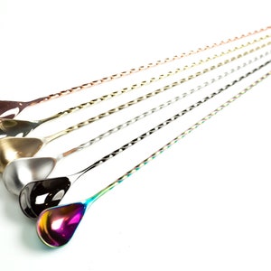 May include: An assortment of bar spoons with twisted handles and teardrop-shaped bowls. The spoons are in various metallic finishes, including copper, silver, and iridescent rainbow colors. The spoons are arranged diagonally against a white background.