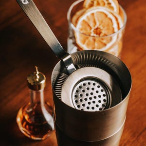 May include: A silver cocktail shaker with a built-in strainer and a silver cocktail muddler. The shaker is on a wooden surface with dried orange slices in a glass behind it.