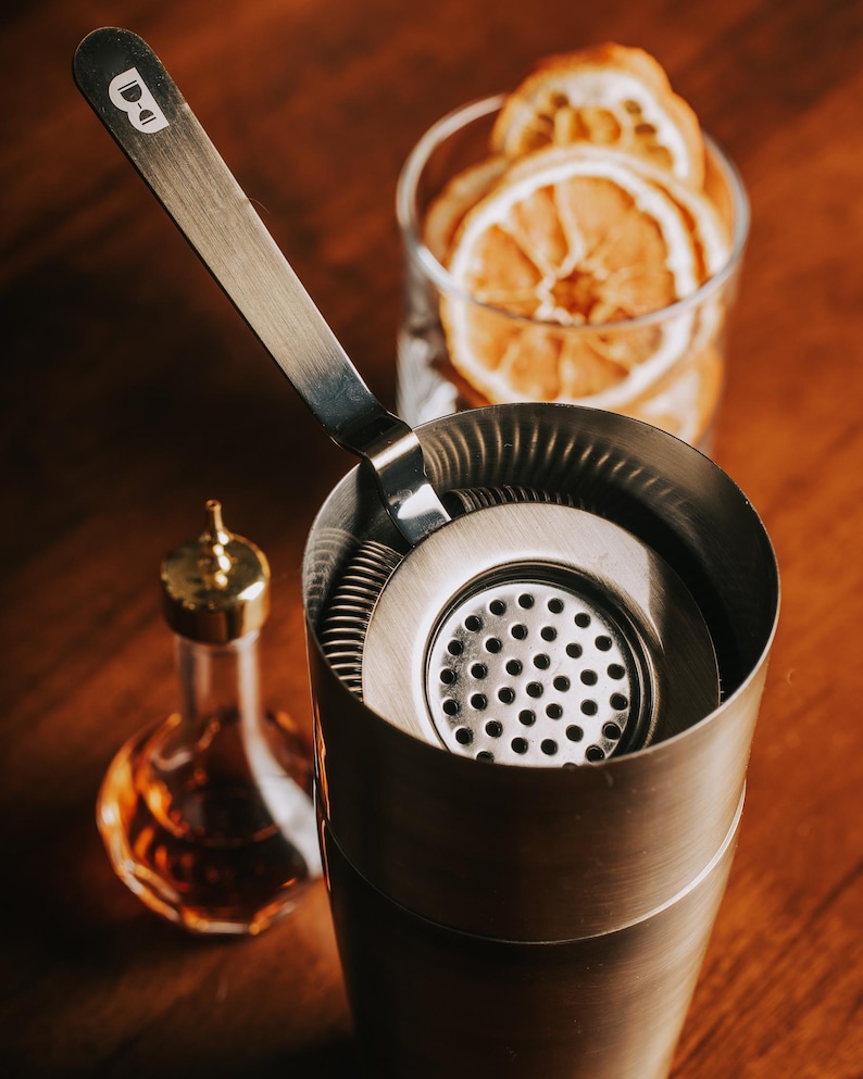 May include: A silver cocktail shaker with a built-in strainer and a silver cocktail muddler. The shaker is on a wooden surface with dried orange slices in a glass behind it.