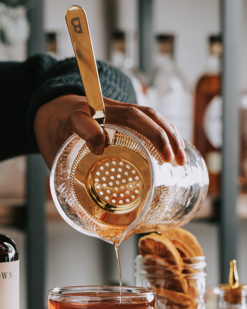 May include: A gold cocktail strainer with a handle is being used to pour a clear liquid from a glass pitcher into a glass. The pitcher has a textured surface and the glass has a clear liquid inside.