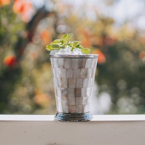 Puede incluir: Un vaso de cóctel decorativo con un patrón de mosaico de baldosas rectangulares de nácar. El borde y la base son de color plateado. El vaso está lleno de hielo y cubierto con hojas de menta fresca. El fondo está borroso, con toques de verde y naranja.