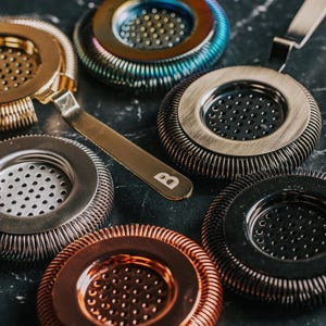 May include: A set of six cocktail strainers in different colors and finishes, including copper, gold, silver, and black. Each strainer has a circular shape with a mesh center and a decorative, textured outer ring.