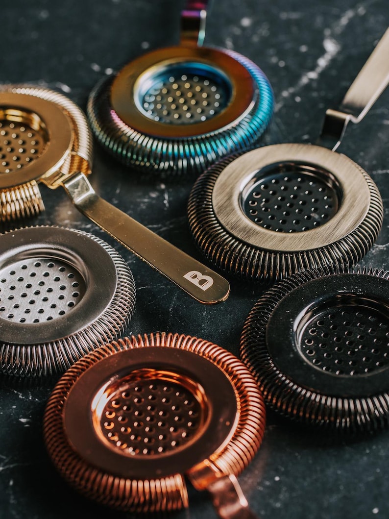 May include: A set of six cocktail strainers in different colors and finishes, including copper, gold, silver, and black. Each strainer has a circular shape with a mesh center and a decorative, textured outer ring.