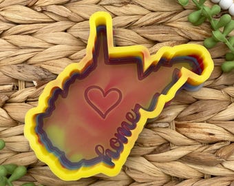 May include: A multi-layered, colorful cookie cutter in the shape of West Virginia. The cutter has a yellow outline, with layers of blue, purple, and red. A red heart is in the center, and the word "home" is written in cursive.